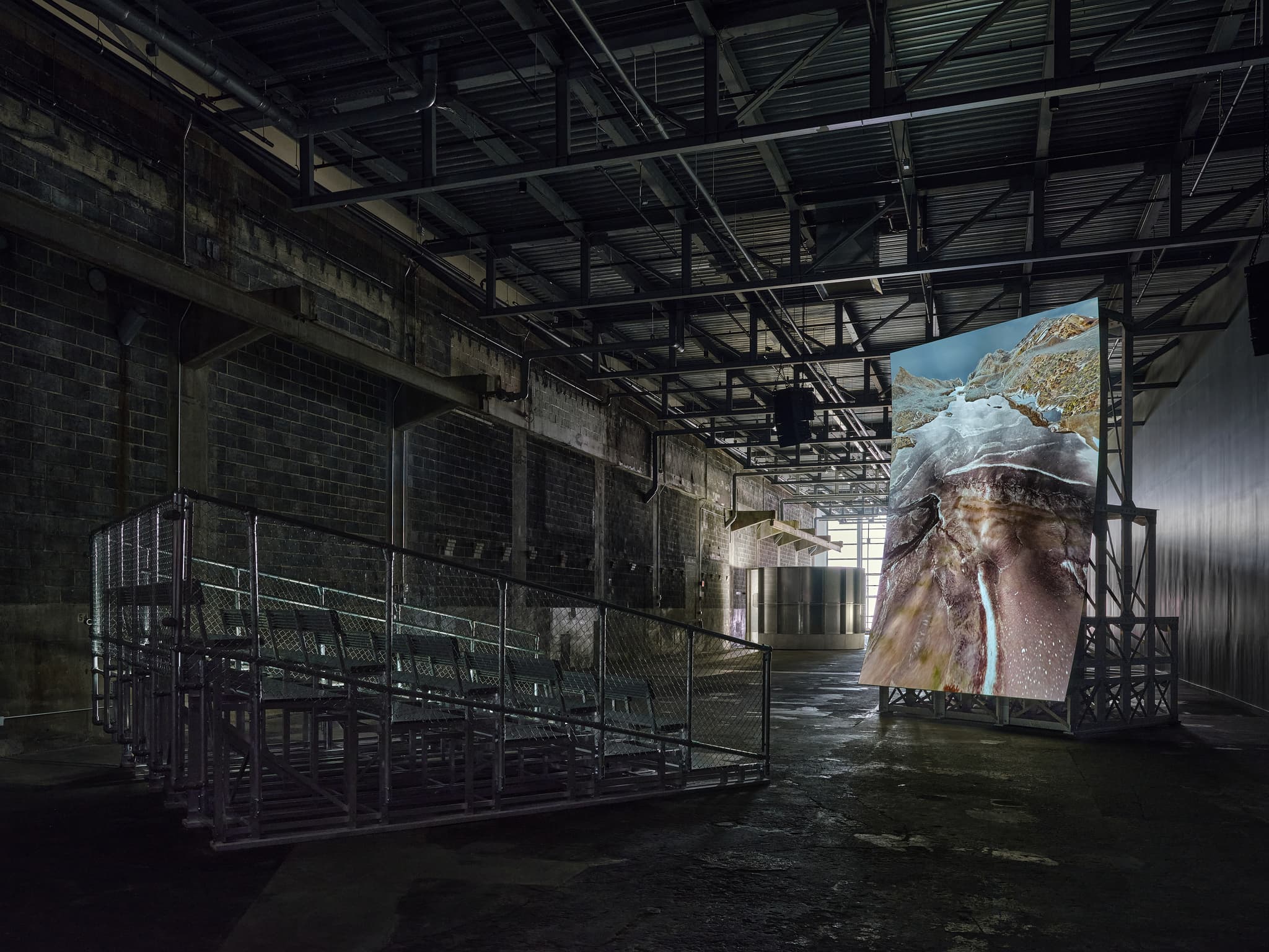 Inside an industrial space, a set of metal bleachers faces a vertical screen displaying an image of rushing water through a cavern.