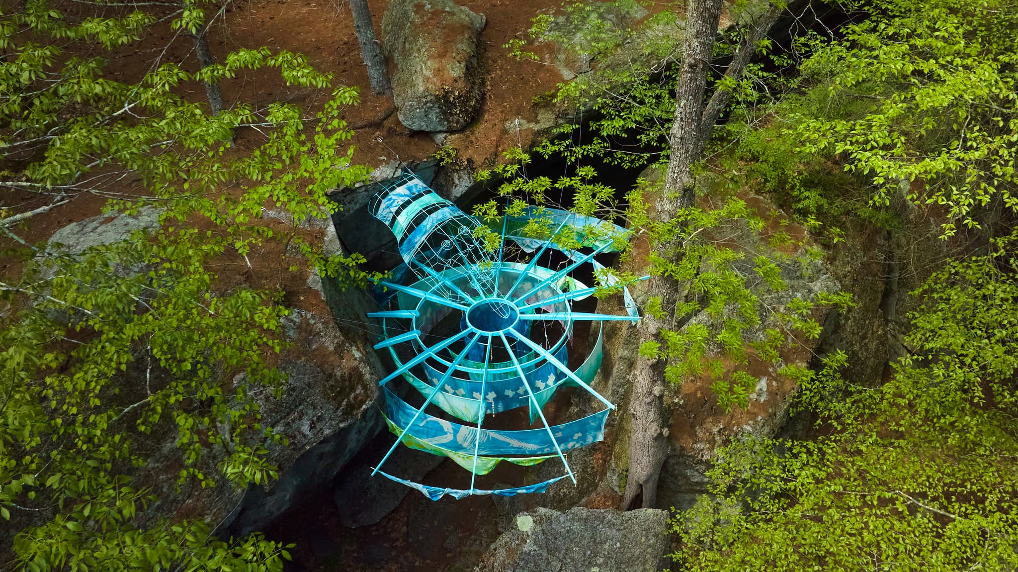 A blue metal sculpture with hanging silks is situated between rocks in a forest.