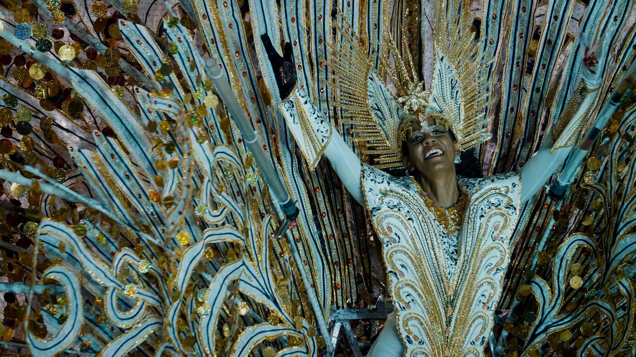 A woman wearing a dazzling costume smiles and raises her arms joyously in the air.